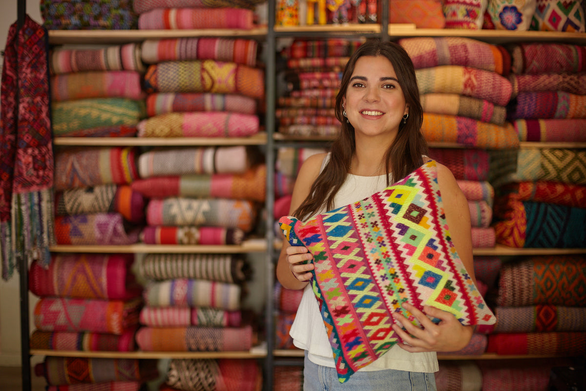Amalia Fuenzalida sosteniendo un cojín en el fondo mantas y aguayos de artesania latinoamericana